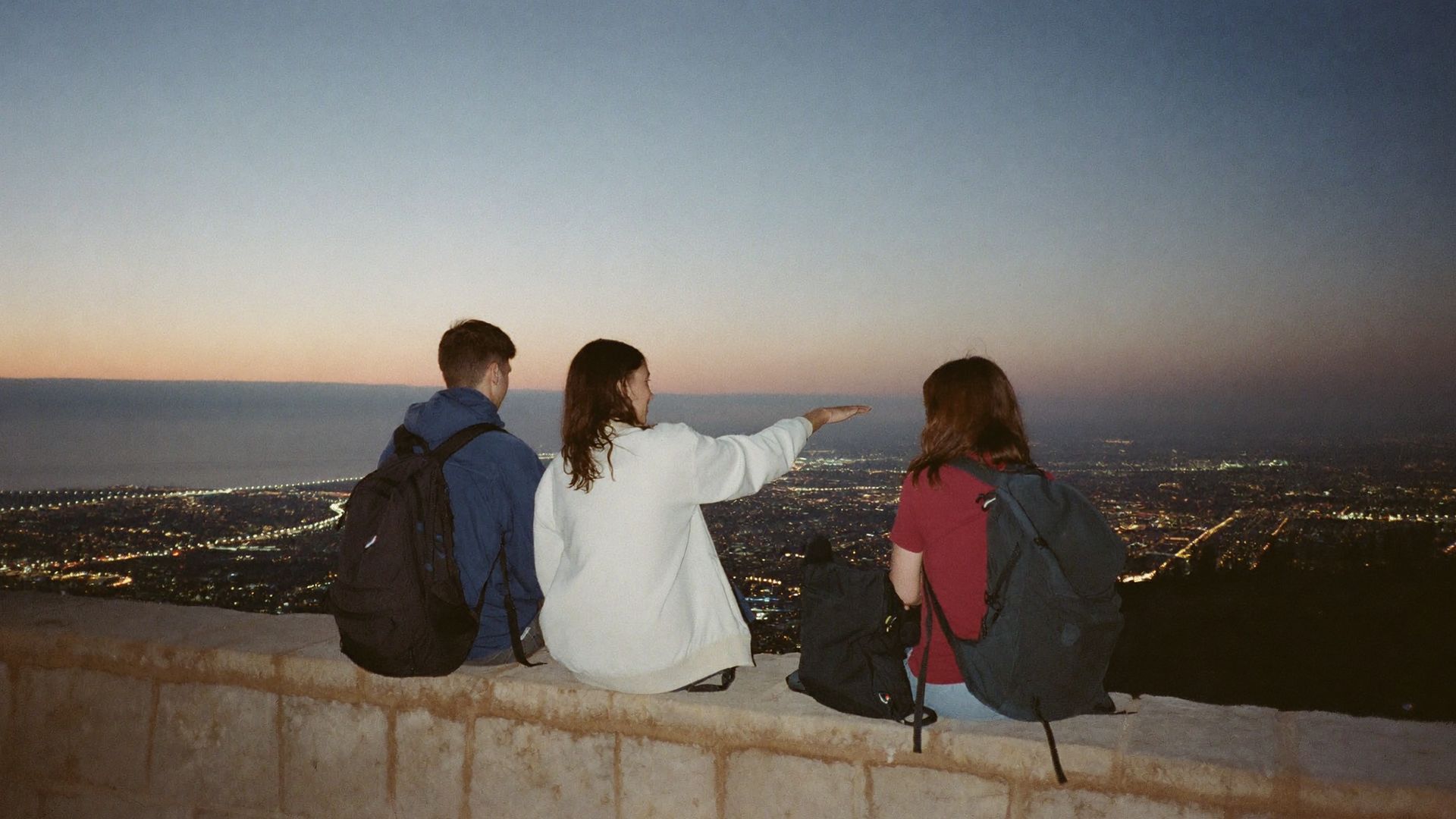 Three friends sitting on a low wall watch as the city comes alive at sunset