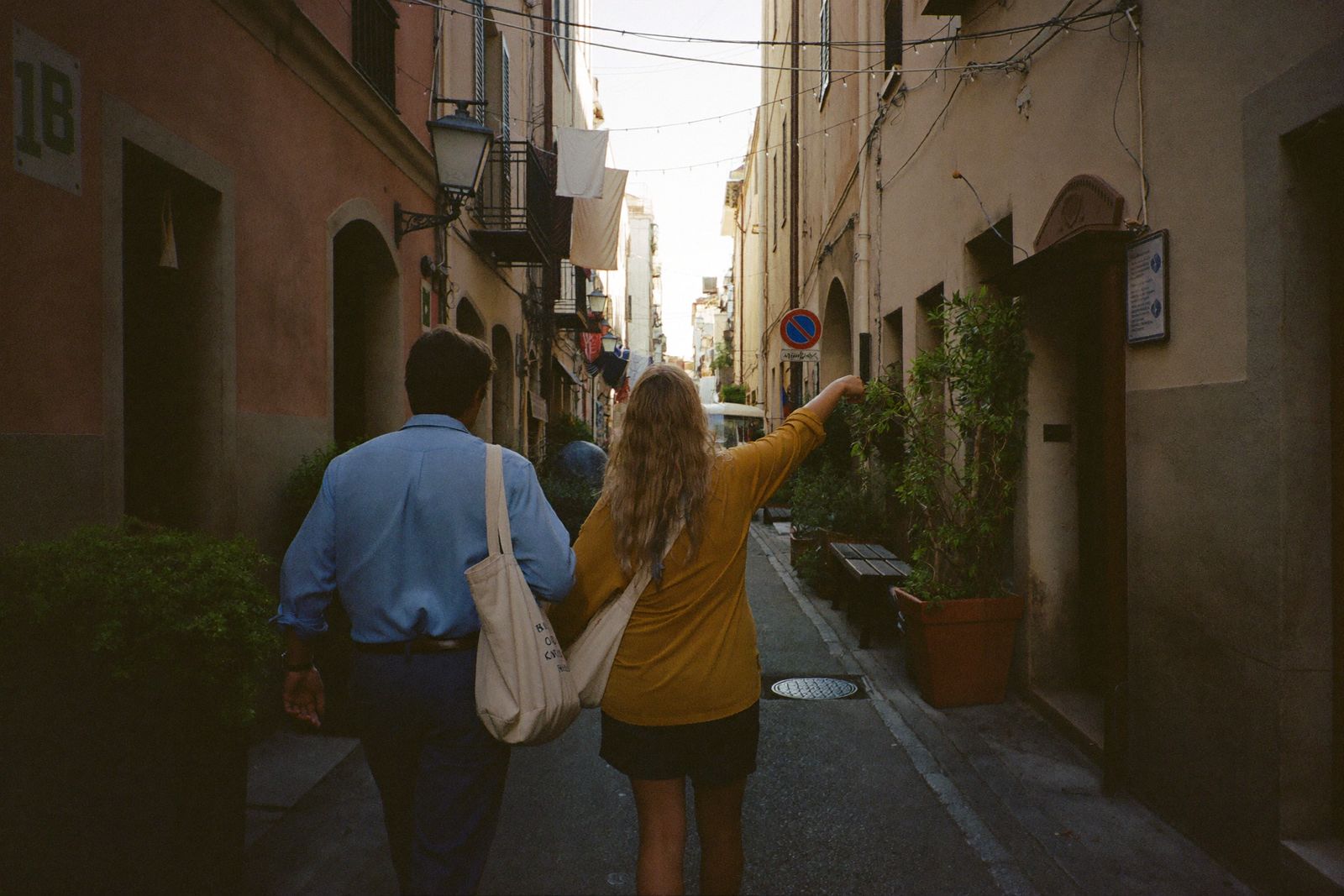 Two friends are walking down a narrow street in a city at sunset: one of them points to a door